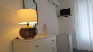 A hotel room with a bedside lamp, a small table with a decorative item, a TV mounted on the wall, and a mini-fridge. - B&B Il Cappero (Lipari)