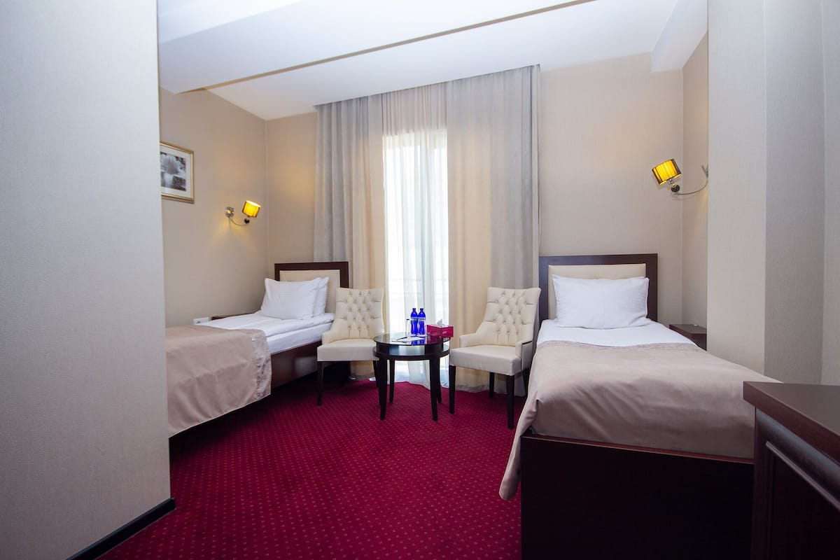 Een hotelkamer met twee eenpersoonsbedden, een klein tafeltje met een stoel en een groot raam met gordijnen.