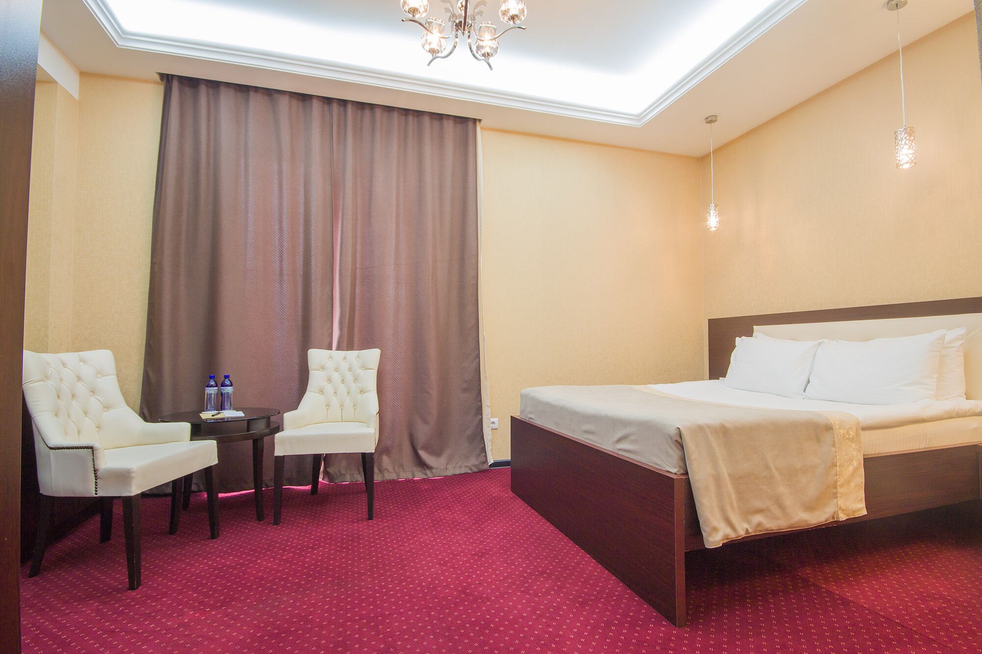A hotel room with a bed, two armchairs, a small table, a chandelier, and a large window with curtains.