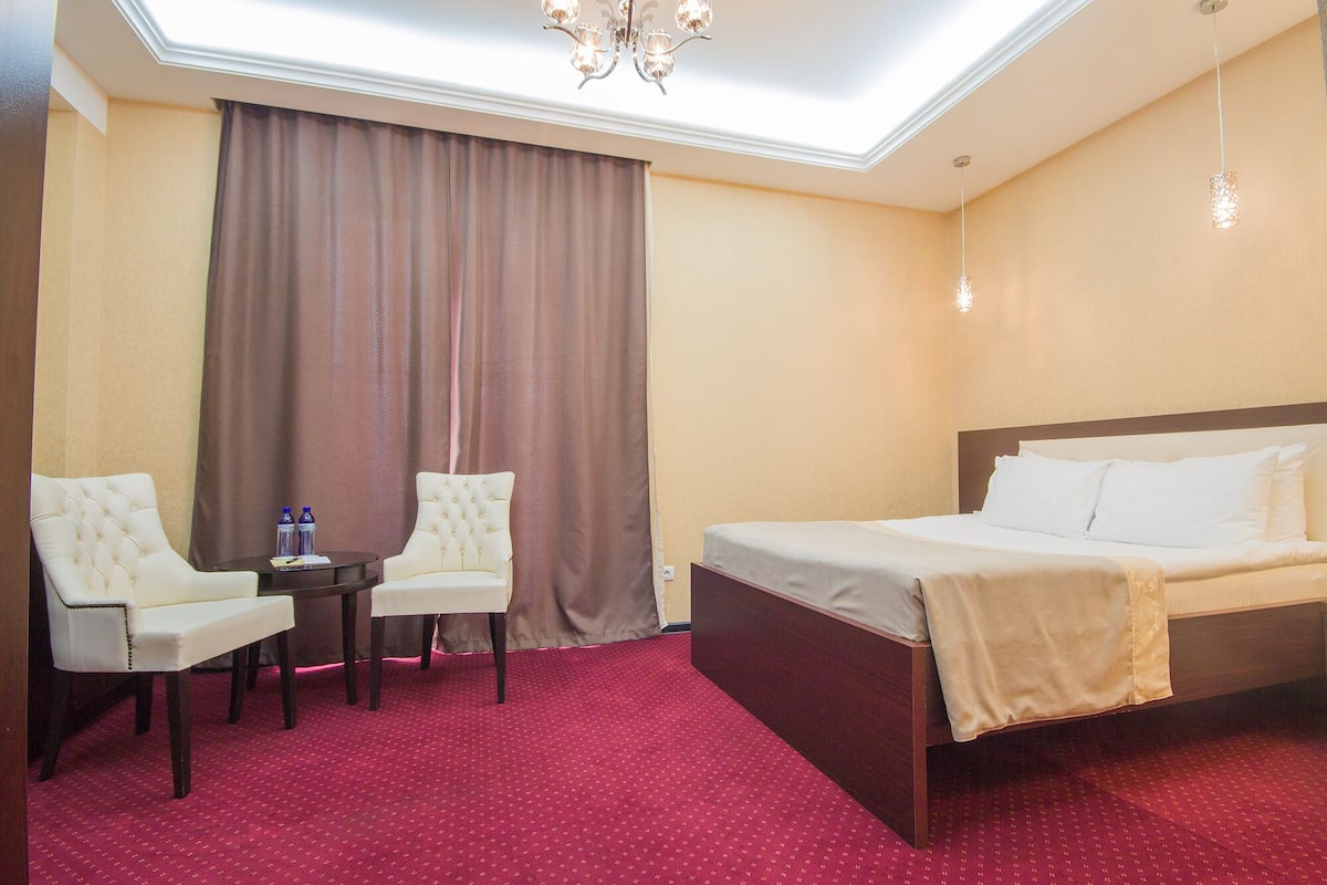 Een hotelkamer met een bed, twee fauteuils, een tafeltje, een kroonluchter en een groot raam met gordijnen.