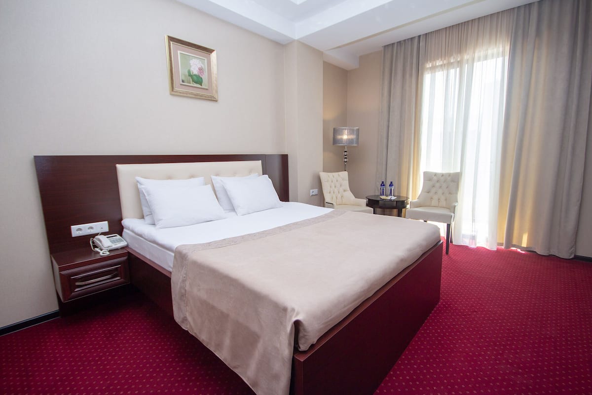 Een hotelkamer met een groot bed, een nachtkastje met telefoon, een stoel, een lamp en een raam met gordijnen.