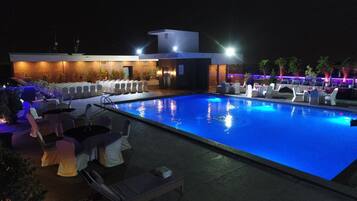 A nighttime pool area with lounge chairs and tables, a modern building with a flat roof, and a pool illuminated by blue lights.