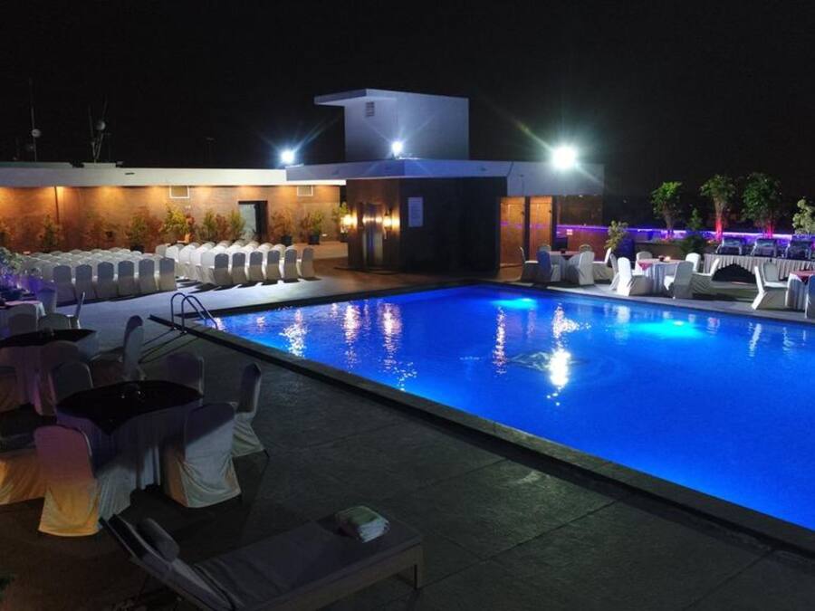 A nighttime pool area with lounge chairs and tables, a modern building with a flat roof, and a pool illuminated by blue lights.