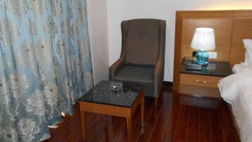 A hotel room with a wooden floor, a small table with a lamp, a chair, and a bed with a headboard.