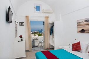 Un dormitorio con cama, televisión, ventana con cortinas, una mesita y vista al mar.
