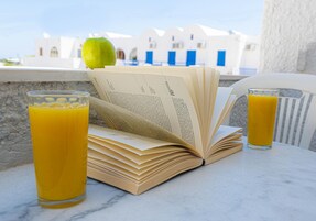 Un libro abierto sobre una mesa, con dos vasos de jugo de naranja, una manzana verde y vistas a edificios blancos con detalles azules.