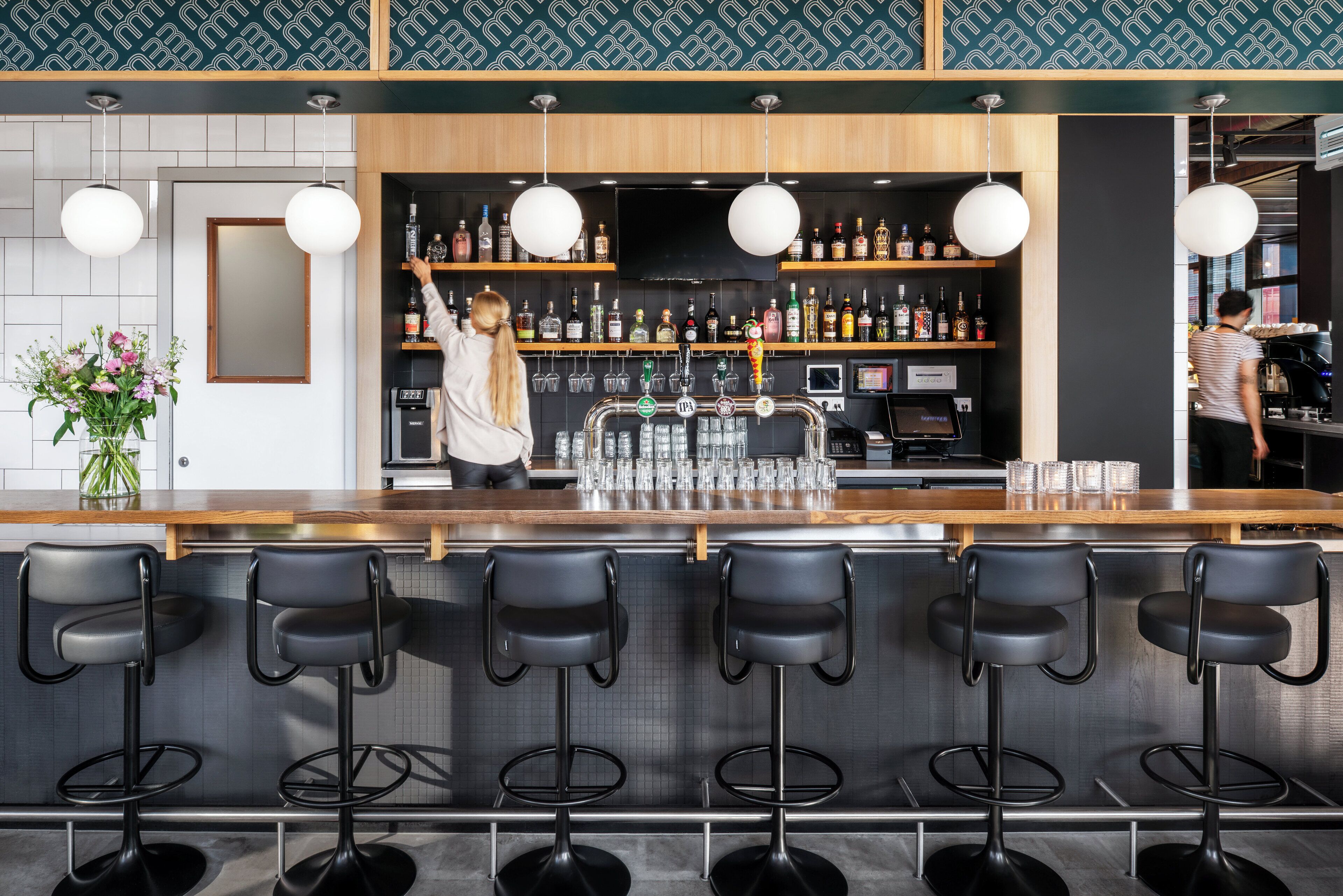 Un espace bar doté d’un comptoir en bois, de tabourets de bar noirs et d’étagères remplies de bouteilles.
