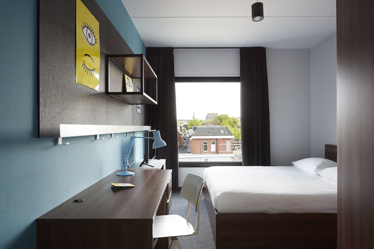 A hotel room with a bed, a desk, a chair, a lamp, a shelf with a picture, and a window with a view of buildings.
