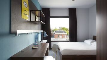 Een hotelkamer met een bed, een bureau, een stoel, een lamp, een plank met een foto en een raam met uitzicht op gebouwen.