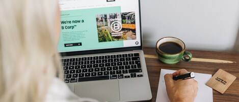 A laptop on a wooden desk with a cup of coffee, a notepad, and a pen.