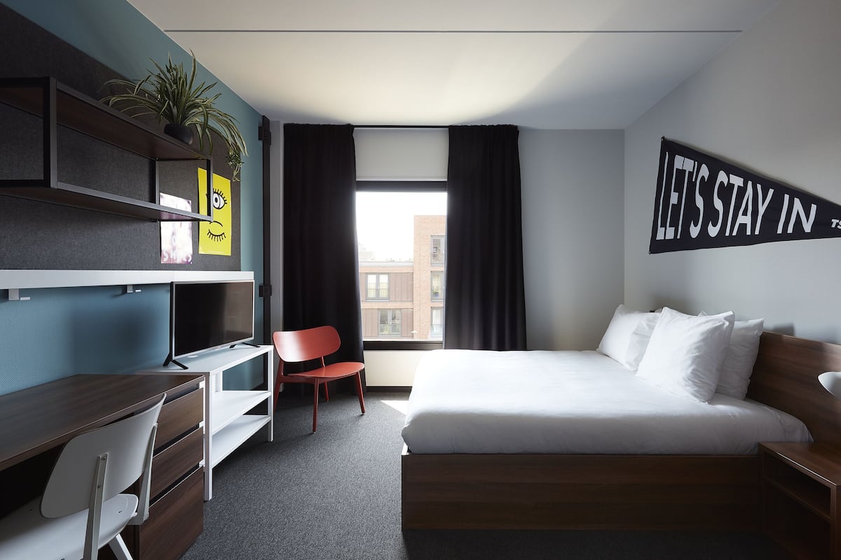 A modern hotel room with a bed, desk, chair, and a large window with curtains.