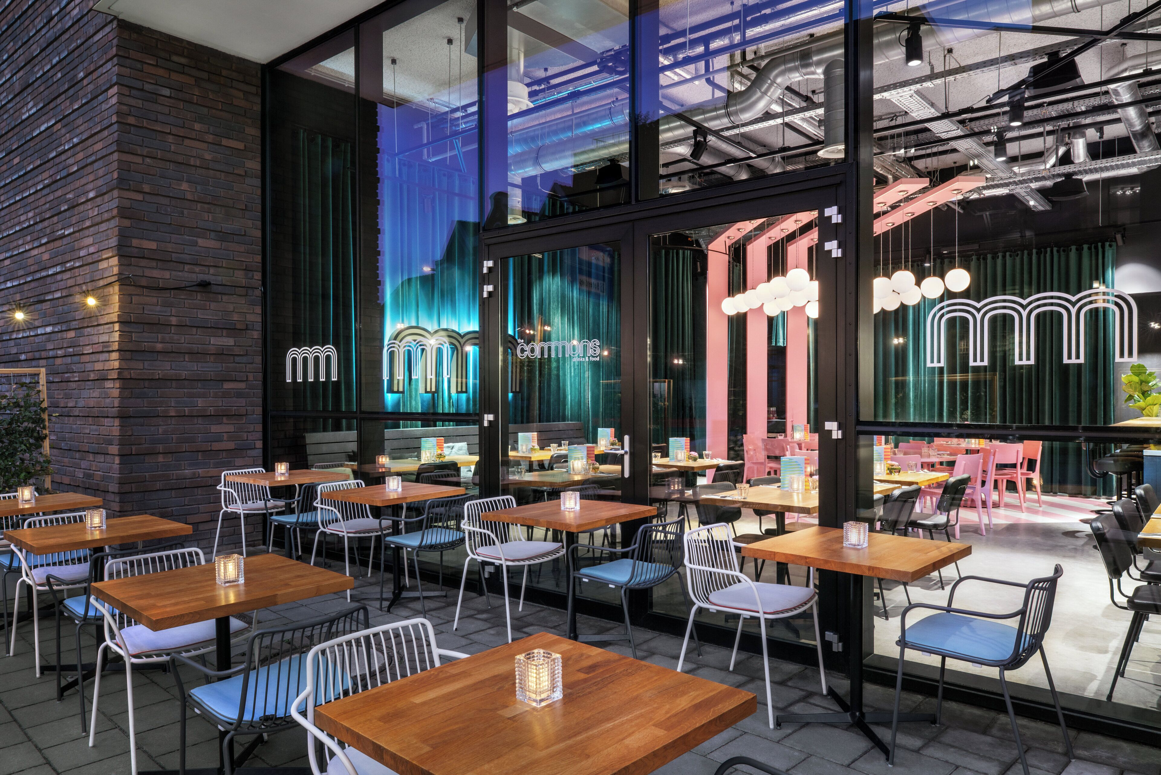 An outdoor cafe with tables and chairs, a brick wall, and large windows with interior lighting.