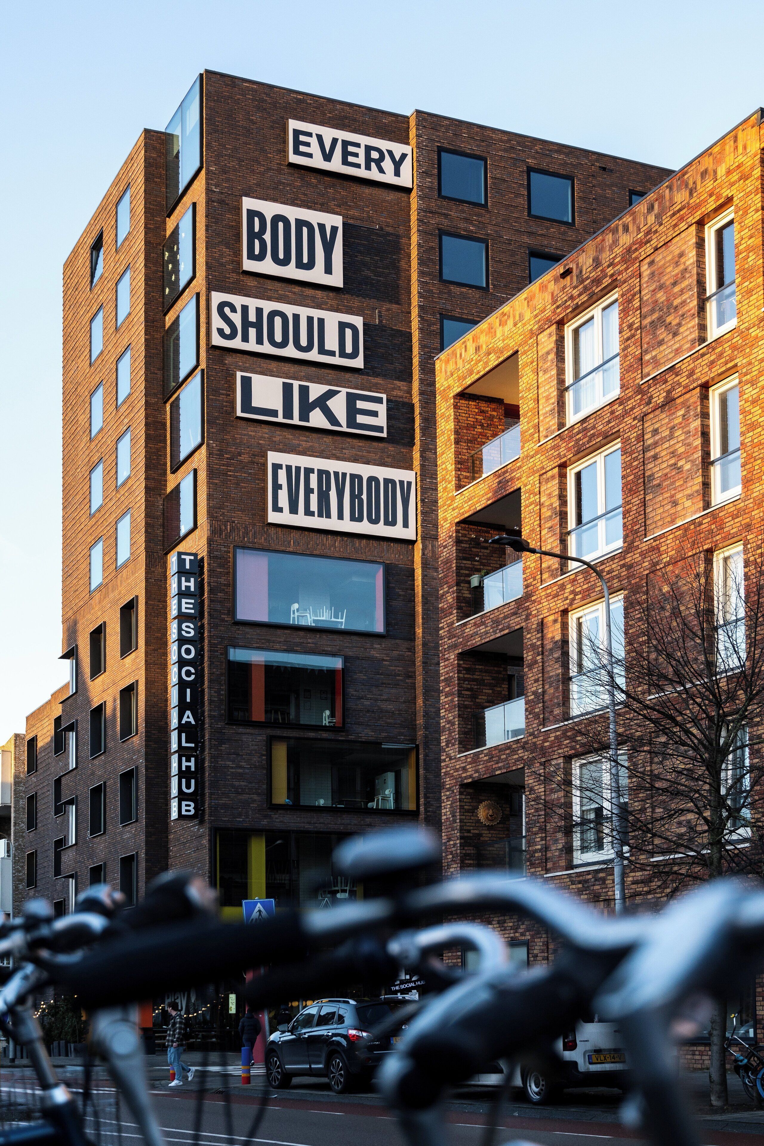 En bygning med flere etager, med et skilt der står: "EVERY BODY SHOULD LIKE EVERYBODY".