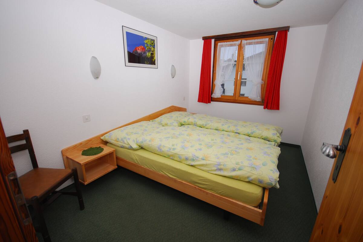 A small, single-bed room with a wooden frame, a green carpet, a wooden chair, a bedside table, a window with red curtains, and a framed picture on the wall.