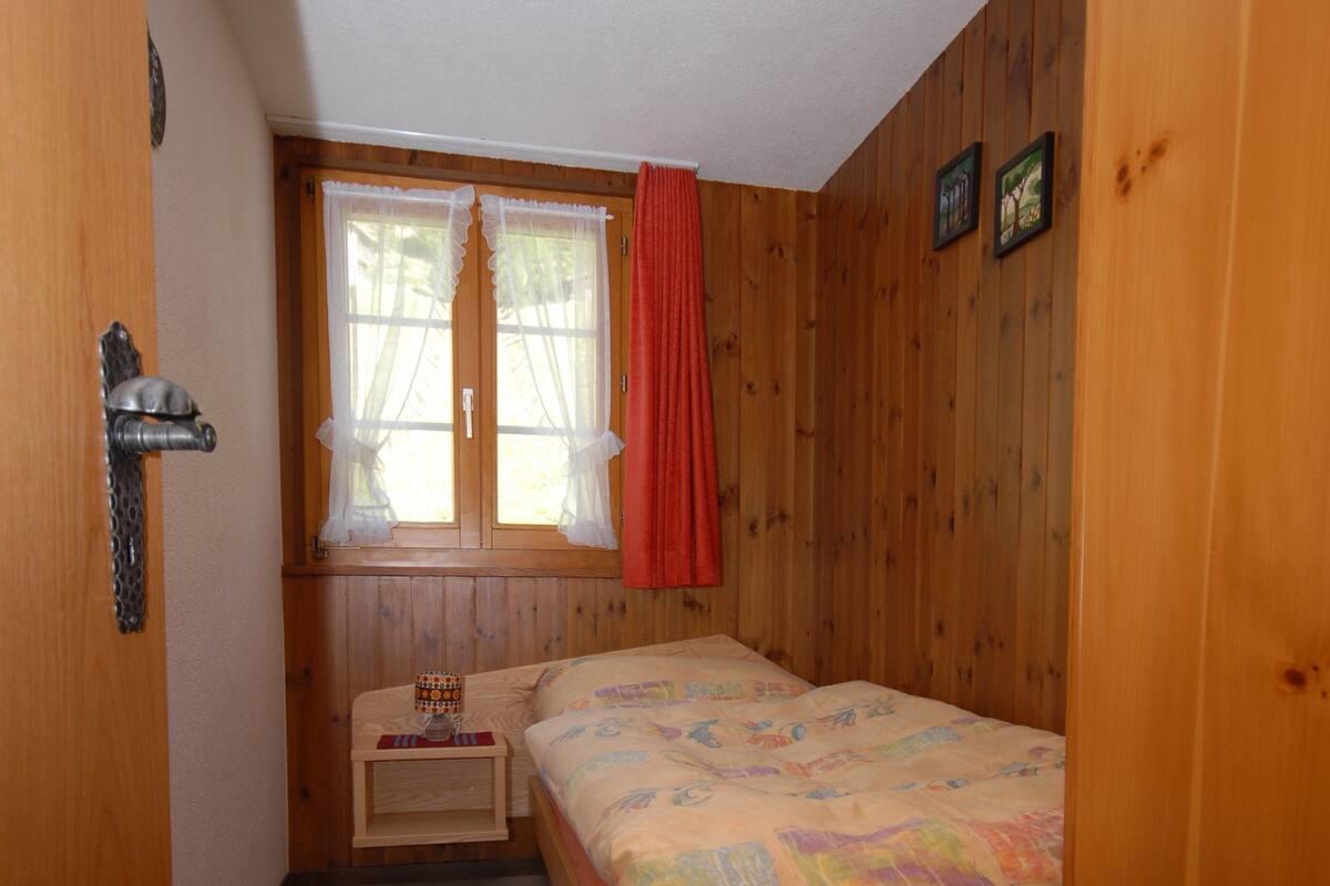 A small, cozy room with a single bed, a nightstand, a window with curtains, and wooden walls.