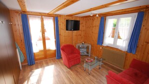 A wooden-paneled room with a TV, two red chairs, a glass coffee table, and a sliding glass door leading to an outdoor area. - Chalet Alpenruh (Saas-Fee)