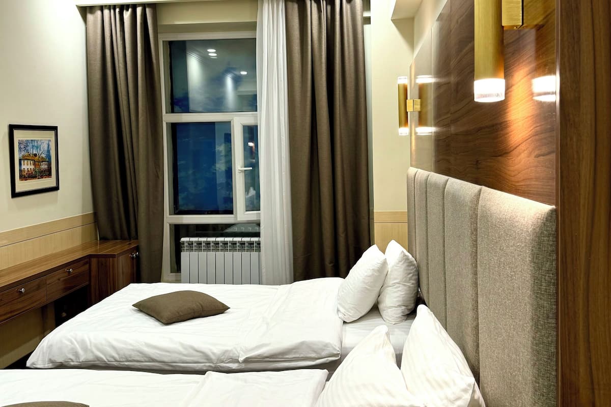 A hotel room with two beds, a large window with curtains, a wooden headboard, and a framed picture on the wall.