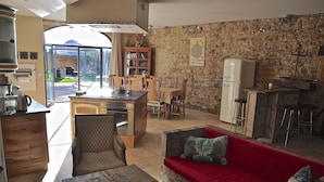 A spacious living area with a red sofa, wooden dining table, and chairs, a kitchen with a stone wall, and a view of the outdoors. - Le Clos du Cher en Beaujolais (Saint-Germain-Nuelles)