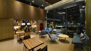 A modern interior with a wooden wall, colorful geometric wall art, a seating area with blue chairs and a sofa, and a view of the street through large windows. - POP! Hotel Pasar Baru Jakarta (Jakarta)