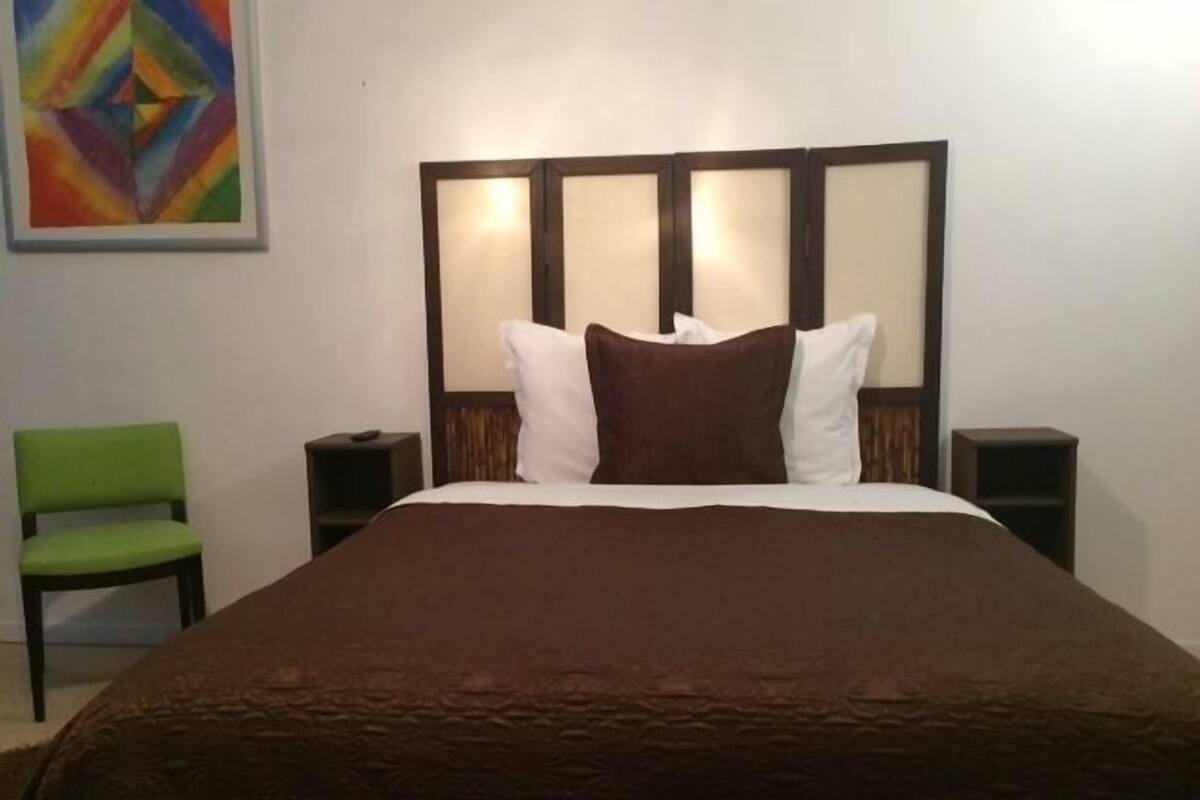 A hotel room with a large bed, a green chair, two bedside tables, and a colorful painting on the wall.