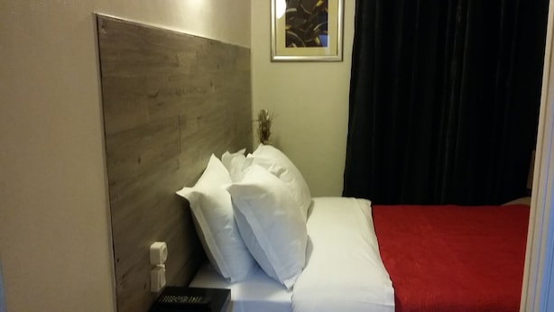 A hotel room with a wooden headboard, a bed with white bedding and a red accent, a remote control on the nightstand, and a framed picture on the wall. - Hôtel Nice Savoie (Nice)