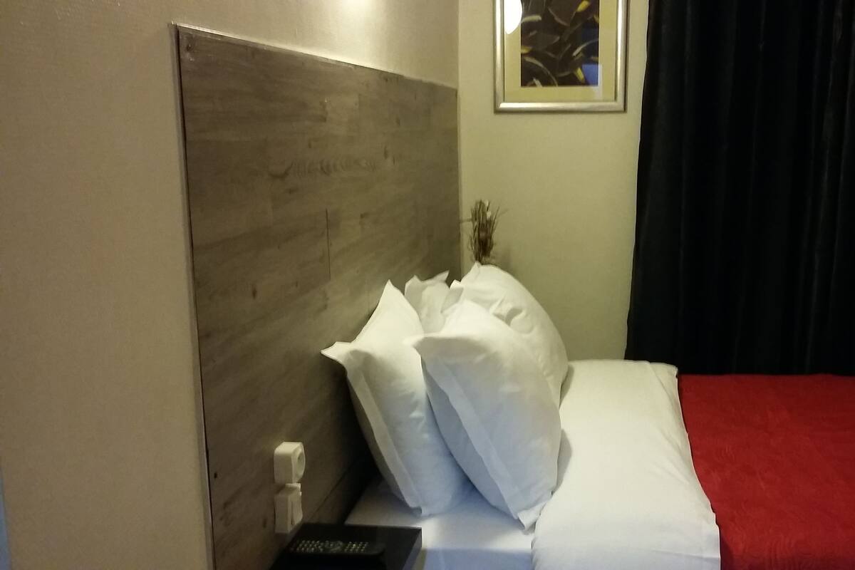 A hotel room with a wooden headboard, a bed with white bedding and a red accent, a remote control on the nightstand, and a framed picture on the wall.