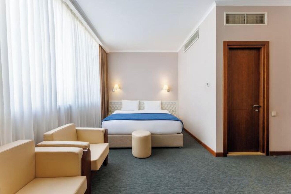A hotel room with a bed, two bedside lamps, a sofa, a chair, and a door.