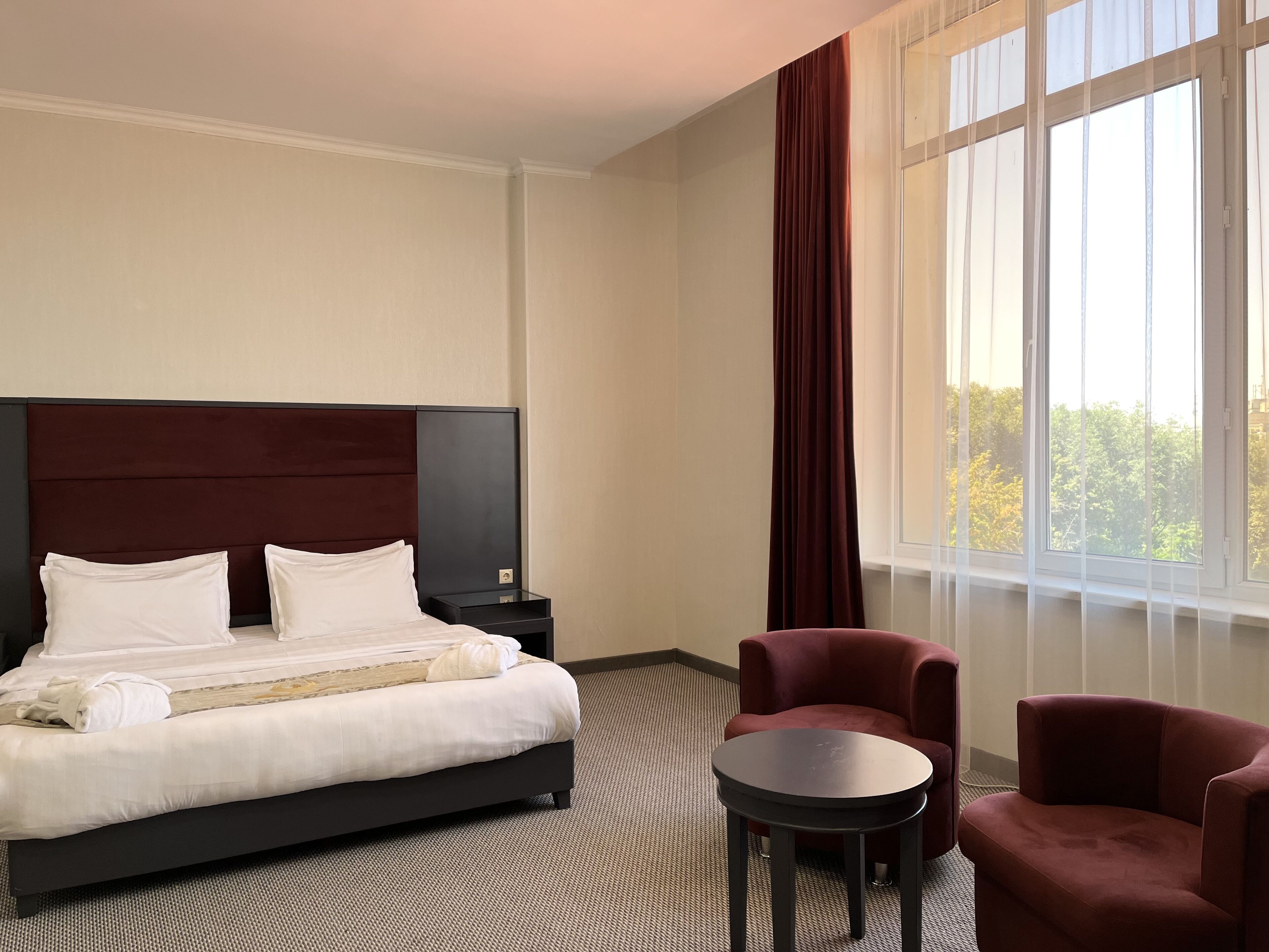 A hotel room with a bed, a nightstand, a round table, two armchairs, and a large window with curtains.