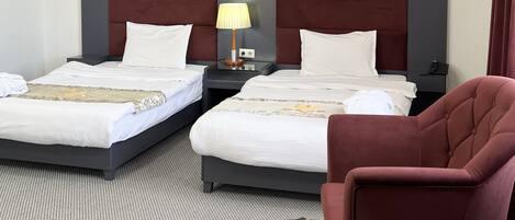 A hotel room with two beds, a nightstand with a lamp, a chair, and a small table with a book.