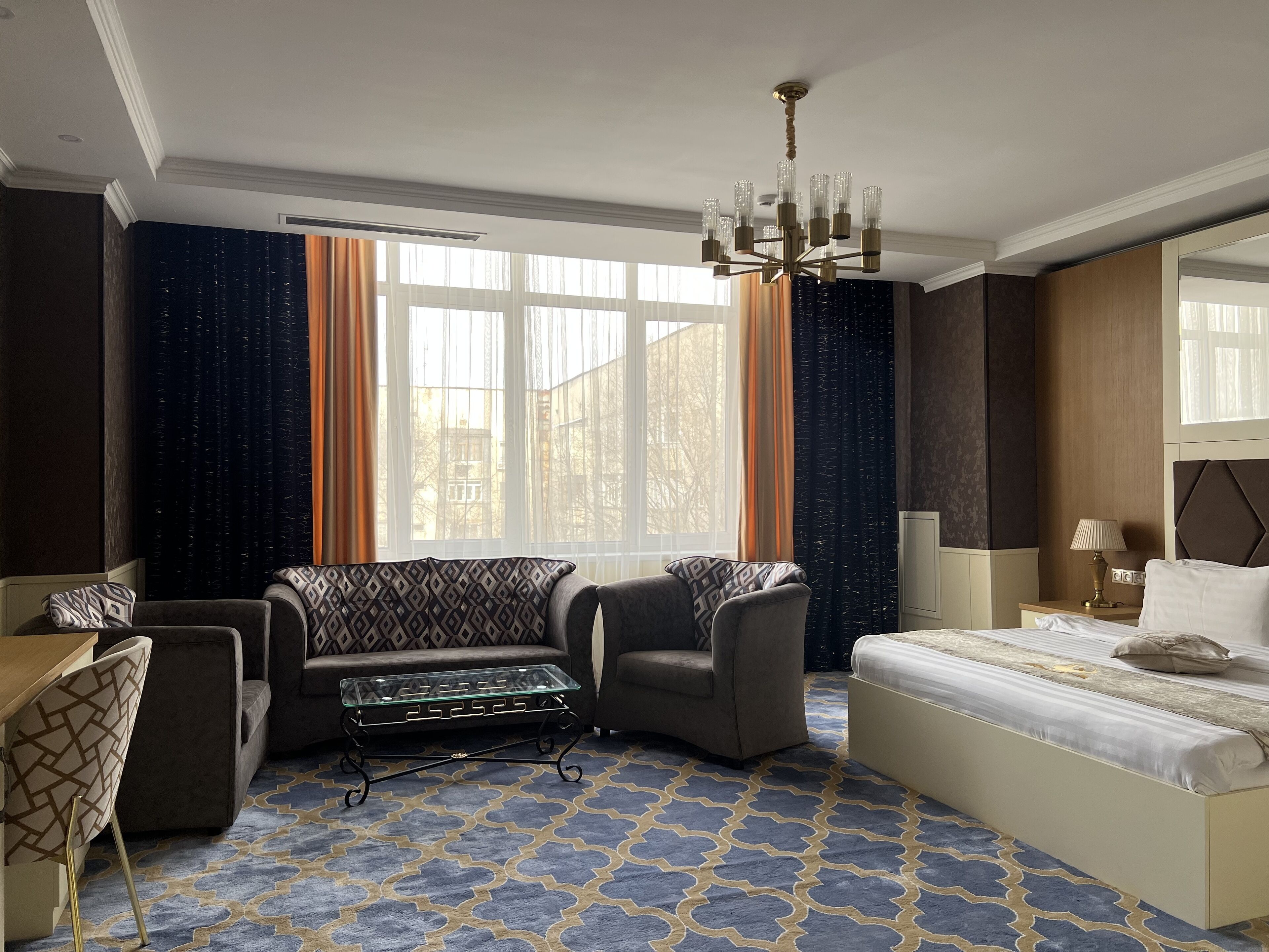 A hotel room with a bed, two armchairs, a small table, a chandelier, and large windows with curtains.