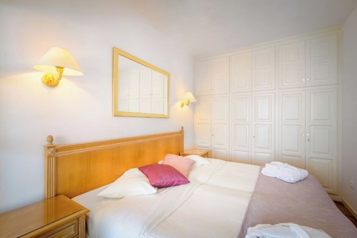 A hotel room with a wooden headboard, a bed with white and pink bedding, two bedside lamps, a mirror, and a large white wardrobe.
