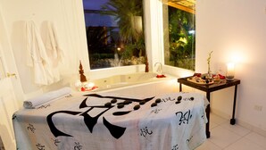 Ein Spa-Zimmer mit Bett, Fenster mit Blick auf Palmen, Badewanne und einem kleinen Tisch mit Kerzen und Steinen.