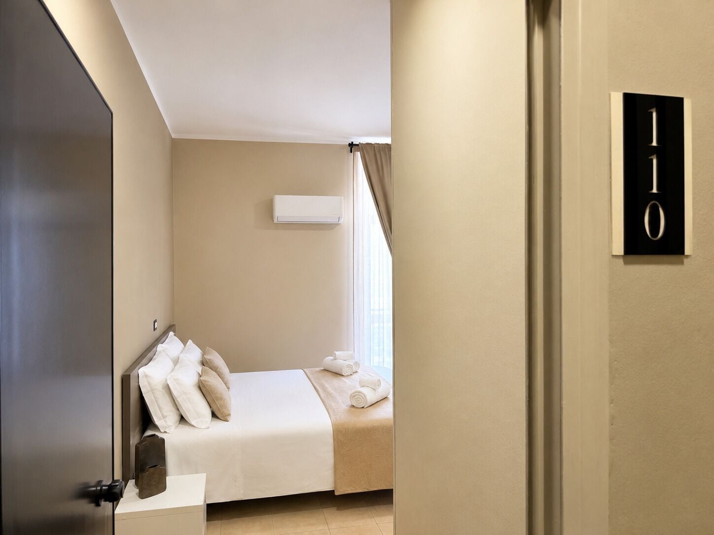 Quarto de hotel com uma cama, janela com cortinas, ar condicionado fixado na parede e porta com sinal de número.