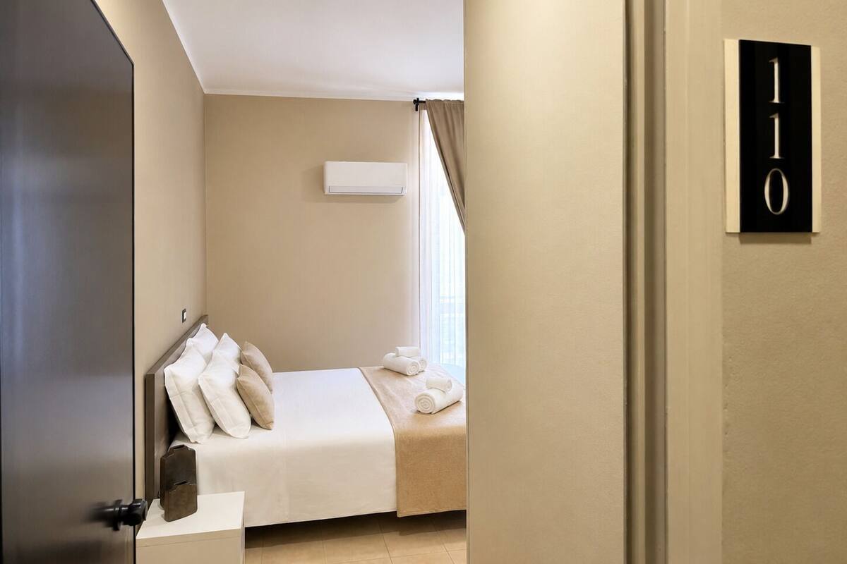 A hotel room with a bed, a window with curtains, a wall-mounted air conditioner, and a door with a number sign.
