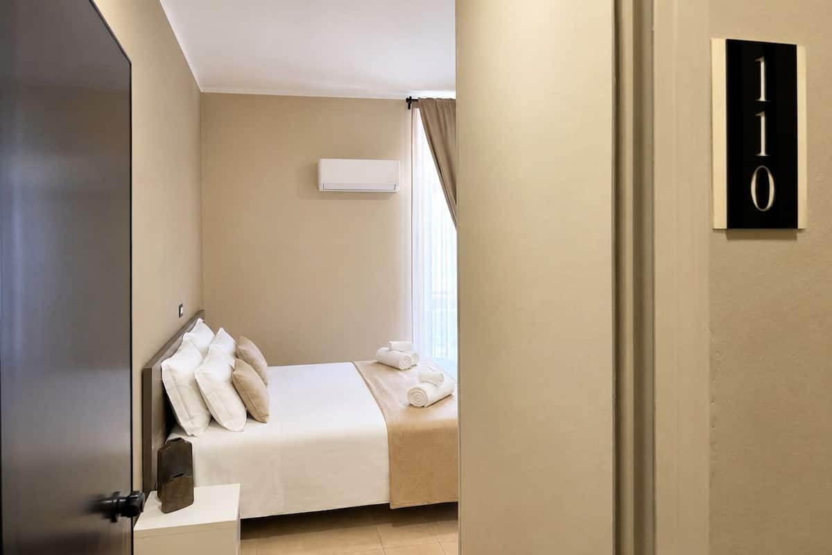 Quarto de hotel com uma cama, janela com cortinas, ar condicionado fixado na parede e porta com sinal de número.