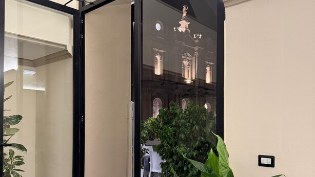 Entrada de hotel con puerta de cristal, felpudo con el nombre del hotel, planta en maceta y vista del interior del hotel a través del cristal.