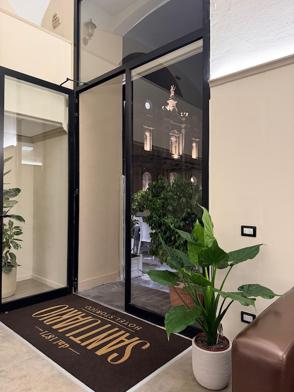 A hotel entrance with a glass door, a doormat with the hotel's name, a potted plant, and a view of the hotel's interior through the glass.
