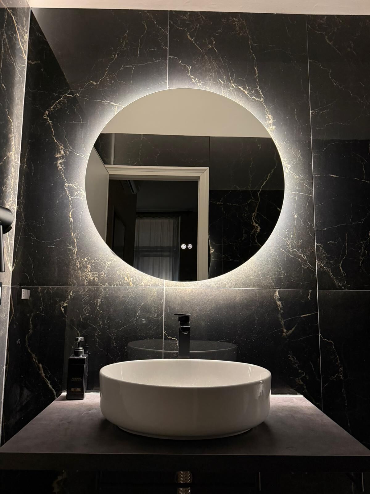 Un baño moderno con un espejo redondo, un lavabo blanco y paredes de mármol oscuro.