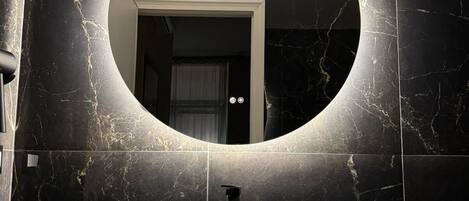 Un baño moderno con un espejo redondo, un lavabo blanco y paredes de mármol oscuro.