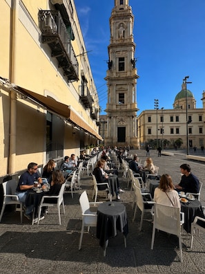 Um café ao ar livre com mesas e cadeiras, um edifício histórico com uma torre de sino e uma cúpula ao fundo.