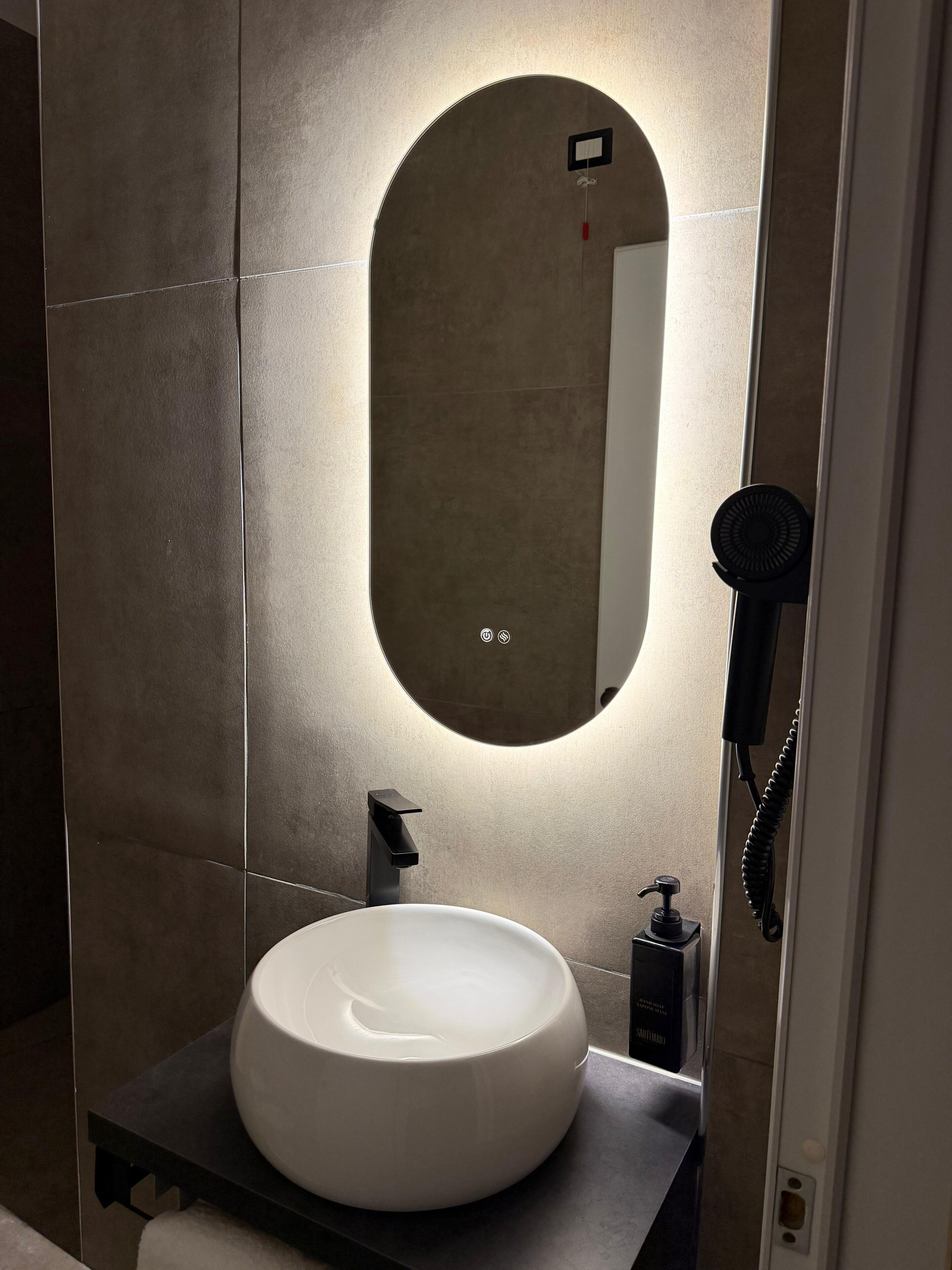 A bathroom with a round white sink, a mirror, and a hairdryer on a wall-mounted shelf.
