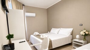 A hotel room with a bed, a desk, a chair, a small table, a plant, and a vase with flowers.