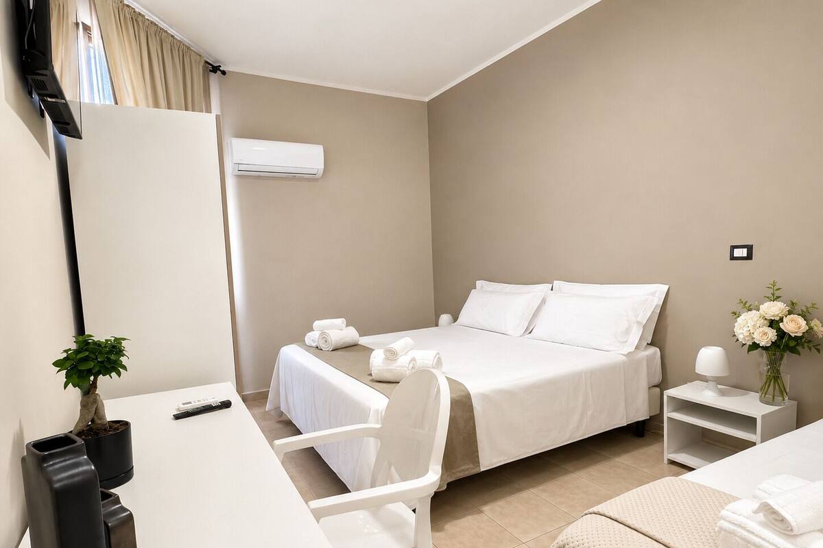 A hotel room with a bed, a desk, a chair, a small table, a plant, and a vase with flowers.