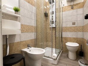 A modern bathroom with a glass-enclosed shower, a pedestal sink, a toilet, and a small shelf with towels and a plant.