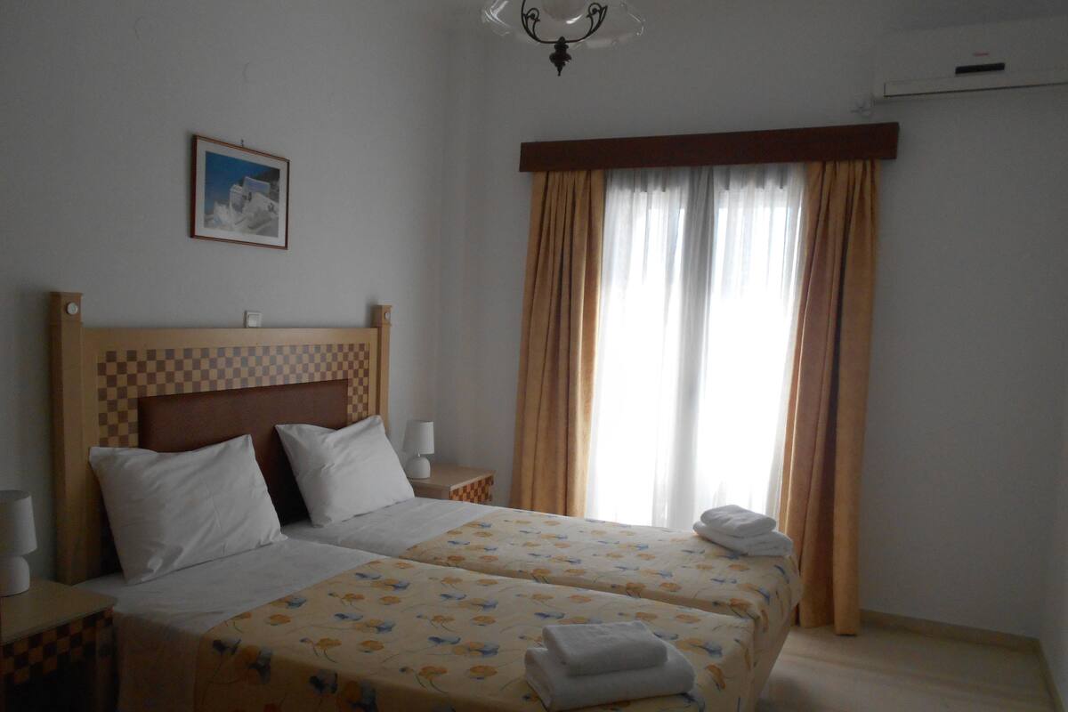 A hotel room with two beds, a checkered headboard, a nightstand with a lamp, a framed picture on the wall, and a window with curtains.