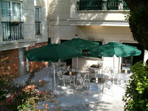 An outdoor seating area with metal chairs and tables, covered by green umbrellas, and a brick building in the background.