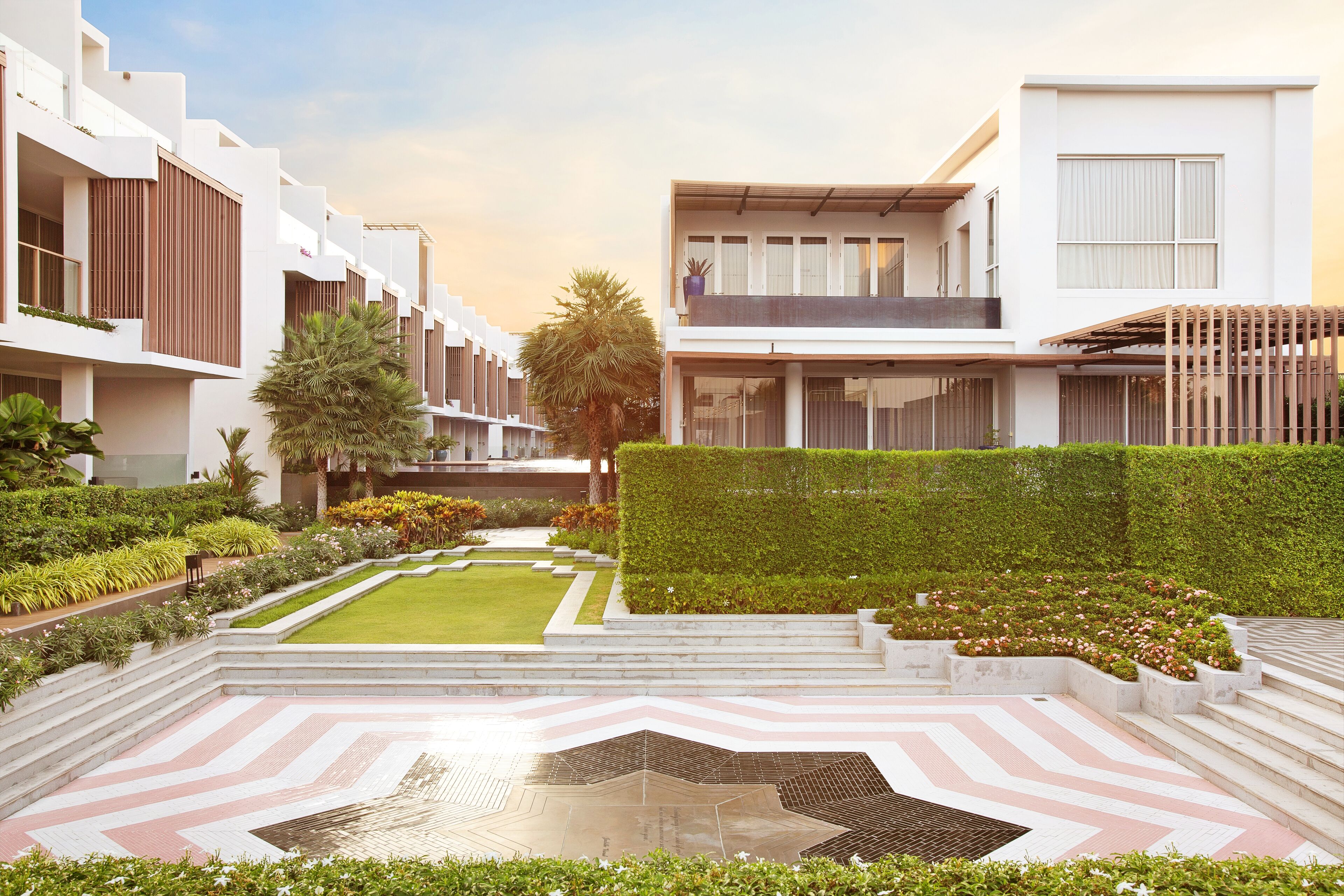 Un edificio moderno con un patio central que presenta un suelo con diseño geométrico, rodeado de vegetación cuidada y un seto perfectamente recortado.