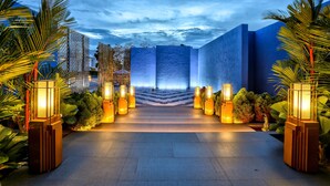 A modern building with a geometric facade, a central entrance, and a well-lit pathway flanked by palm trees and illuminated lanterns.