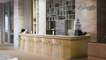 Un área de bar moderna con mostrador de madera, taburetes de metal blancos y estantes con diversas botellas y cristalería.