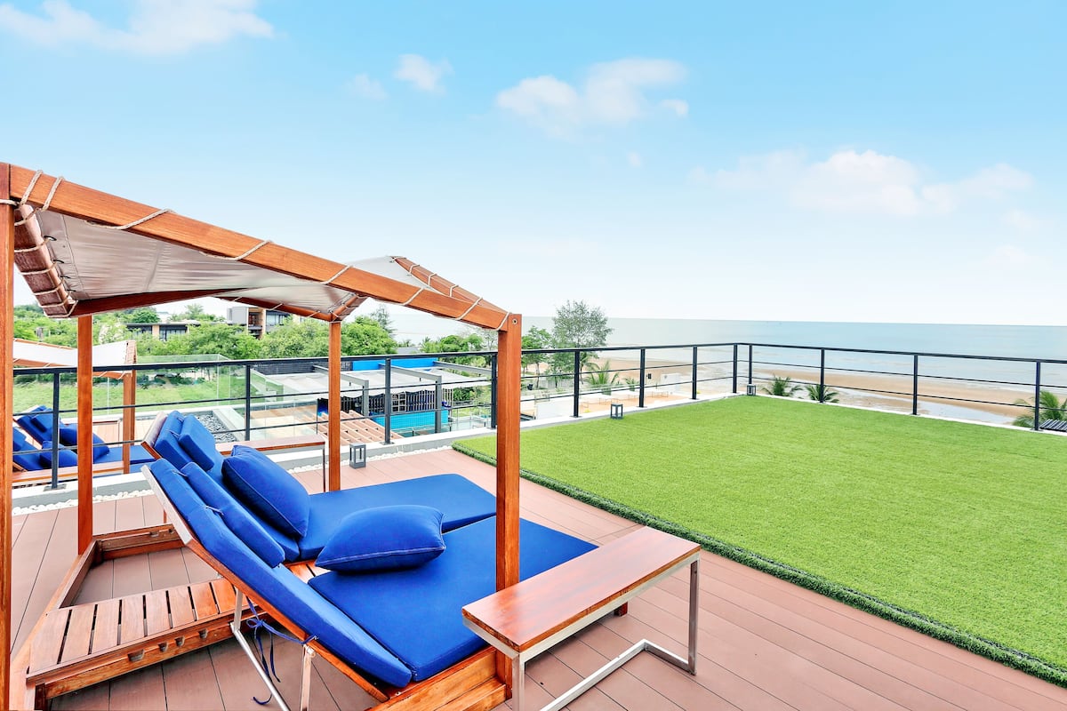 Eine Holzterrasse mit blauem Liegestuhl und einem kleinen Tisch, mit Blick auf einen Pool und eine Rasenfläche mit Meerblick.